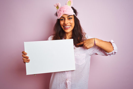 Young woman wearing pajama and sleep mask holding banner over isolated pink background with surprise face pointing finger to himself