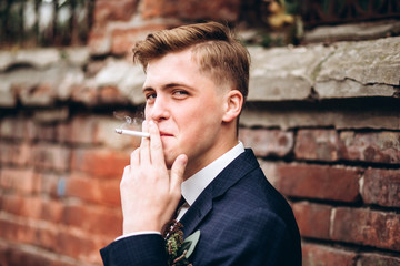 brutal groom in a suit and tie smokes a cigarette. The blond guy is walking around the old town. smoking, bad habits. Young male businessman on brick wall background. Portert of a smiling guy.
