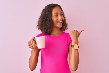 Young brazilian woman drinking cup of coffee standing over isolated pink background pointing and showing with thumb up to the side with happy face smiling