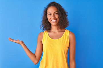 Fototapeta na wymiar Young brazilian woman wearing yellow t-shirt standing over isolated blue background smiling cheerful presenting and pointing with palm of hand looking at the camera.
