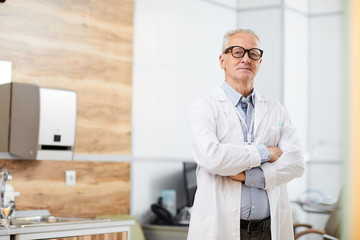 Portrait of confident senior doctor looking at camera while posing standing in office of modern clinic, copy space