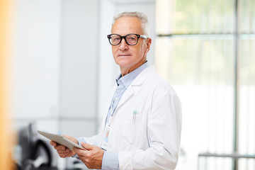 Waist up portrait of senior doctor looking at camera while using digital tablet standing in office...