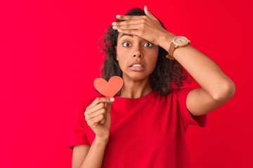 Young brazilian woman holding paper heart standing over isolated red background stressed with hand on head, shocked with shame and surprise face, angry and frustrated. Fear and upset for mistake.