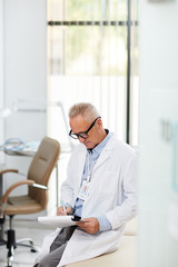 Naklejka na ściany i meble High angle portrait of senior doctor writing on clipboard during consultation in office of modern clinic, copy space