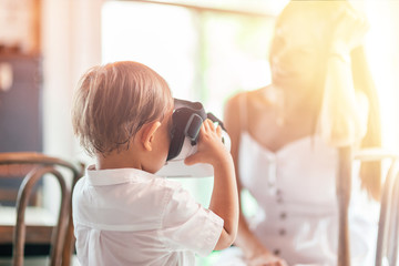 Portrait of 4-year-old boy in white shirt having fun with virtual reality headset indoors at home. His mother sitting in the background. Generation Z concept. Horizontal shot. Selective focus