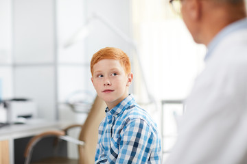 Fototapeta premium Portrait of cute red haired kid looking at senior doctor during consultation in child healthcare clinic, copy space