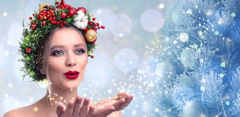 Beautiful young woman with Christmas wreath blowing magical snowy dust on blurred background. Bokeh effect