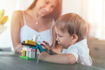 Obraz na płótnie Canvas Portrait of young woman and 4-year-old boy having fun. Cute kid is playing with colorful plastic blocks, while his mother looking at him with a smile. Mom and son concept. Horizontal shot