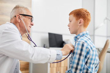 Side view portrait of senior doctor using stethoscope listening to heartbeat and breathing of cute red haired boy during consultation in child healthcare clinic, copy space