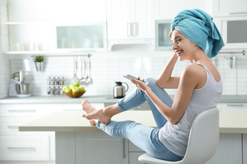 Fototapeta na wymiar Pretty young woman with towel on head using smartphone in kitchen
