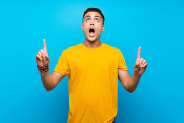 Young man with yellow shirt over isolated blue background surprised and pointing up