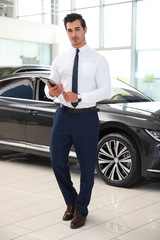 Young salesman with tablet near car in modern dealership