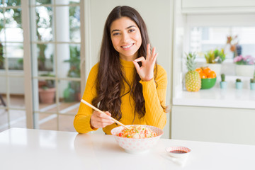 Young woman eating a bowl of Asian rice using chopsticks doing ok sign with fingers, excellent...