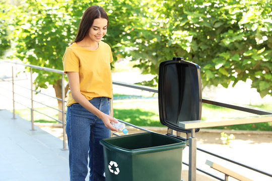 Young woman throwing plastic bottle into recycling bin outdoors