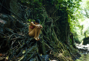 beautiful woman at tropical enchanted forest . Young attractive girl in bikini sitting vulnerable and mysterious on tree trunk at tropical paradise jungle with magical feeling