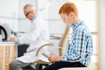 Fototapeta premium Side view portrait of red haired teenage boy using digital tablet in waiting room at doctors office, copy space
