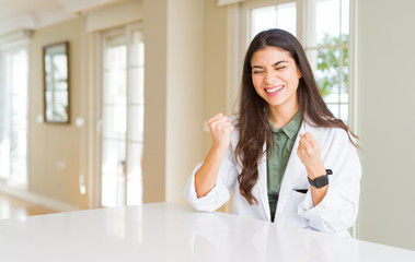 Young woman wearing medical coat at the clinic as therapist or doctor very happy and excited doing winner gesture with arms raised, smiling and screaming for success. Celebration concept.