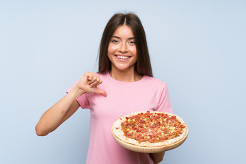 Pretty young girl holding a pizza over isolated blue wall proud and self-satisfied