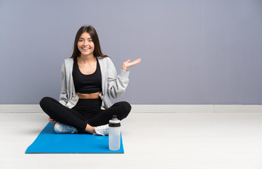 Young sport woman sitting on the floor with mat holding copyspace imaginary on the palm