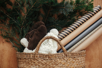 Rustic basket with fir branches,festive wrapping paper, cozy gloves and hat on rustic wooden background. Flat lay. Winter holiday preparations. Slow living lifestyle.