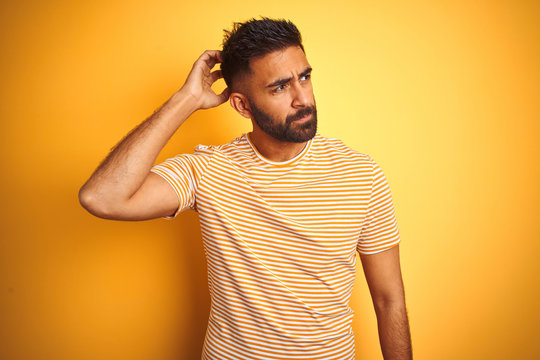 Young indian man wearing t-shirt standing over isolated yellow background confuse and wondering about question. Uncertain with doubt, thinking with hand on head. Pensive concept.