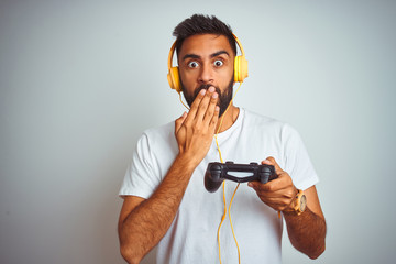 Arab indian gamer man playing video game using headphones over isolated white background cover mouth with hand shocked with shame for mistake, expression of fear, scared in silence, secret concept © Krakenimages.com