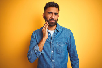 Fototapeta premium Young indian man wearing denim shirt standing over isolated yellow background touching mouth with hand with painful expression because of toothache or dental illness on teeth. Dentist concept.