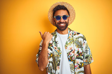 Indian man on vacation wearing floral shirt hat sunglasses over isolated yellow background smiling with happy face looking and pointing to the side with thumb up.