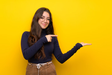 Teenager girl over isolated yellow wall holding copyspace imaginary on the palm to insert an ad