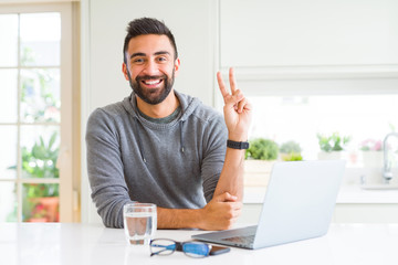 Handsome hispanic man working using computer laptop smiling with happy face winking at the camera doing victory sign. Number two.