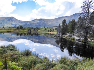 Fototapeta premium View on an Alpine lake, with tall trees growing on the sides. The lake has dark blue color. Calm surface of the lake lets the sky and the surrounding trees reflect in its surface. Boulders on the side