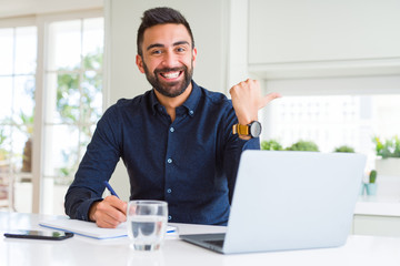 Handsome hispanic man working using computer and writing on a paper pointing and showing with thumb up to the side with happy face smiling