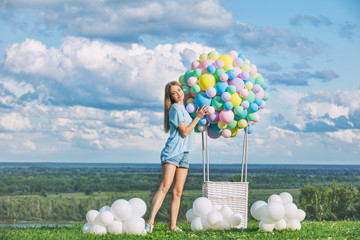 Young adult beautiful blonde girl happy on green grass with balloon airship on blue sky background with clouds