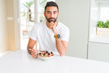 Handsome hispanic man eating healthy breakfast in the morning at home serious face thinking about question, very confused idea