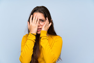 Teenager girl over isolated blue wall covering eyes and looking through fingers