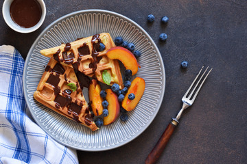 Vanilla Belgian waffles with chocolate sauce, peaches and berries in a plate on the kitchen table.