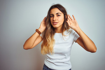 Fototapeta premium Young beautiful woman wearing casual white t-shirt over isolated background Trying to hear both hands on ear gesture, curious for gossip. Hearing problem, deaf