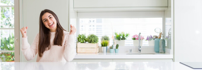 Wide angle picture of beautiful young woman sitting on white table at home celebrating surprised and amazed for success with arms raised and open eyes. Winner concept.