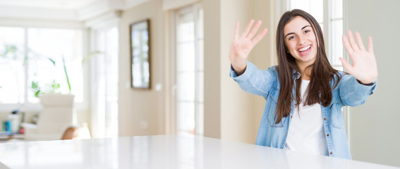 Wide angle picture of beautiful young woman sitting on white table at home showing and pointing up with fingers number nine while smiling confident and happy.