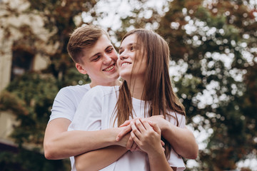 Fototapeta premium Happy young couple walks the streets of the city and hold hands. guy and girl in white t-shirts and jeans outdoors. Teenagers cuddling against the backdrop of an autumn tree. Couple close-up portrait