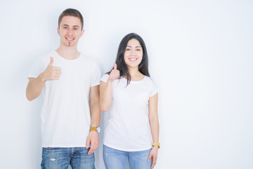 Young beautiful couple wearing casual t-shirt standing over isolated white background doing happy thumbs up gesture with hand. Approving expression looking at the camera with showing success.