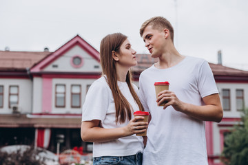 Happy young couple walks the streets of the city and drinks coffee from a cardboard cup. guy and girl in white t-shirts and jeans outdoors. Teenagers cuddle against the backdrop of the cityscape.