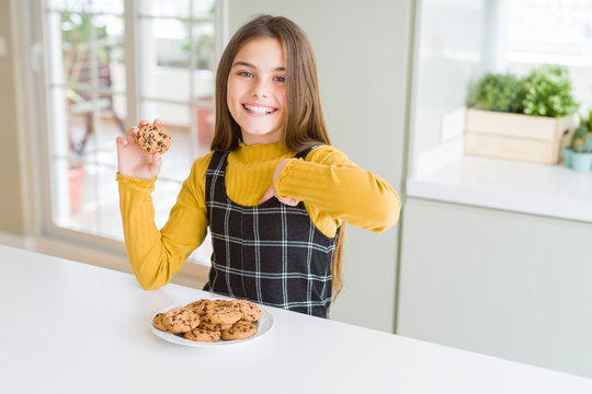Beautiful young girl kid eating chocolate chips cookies with surprise face pointing finger to himself