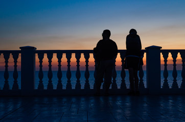 Husband and wife stand and watch the sunset by the sea near the fence of balusters