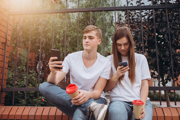 A young couple drinks coffee from cardboard cups and looks at smartphones. Teenagers guy and girl are sitting outdoors. Modern technologies. The problem of communication in modern society.