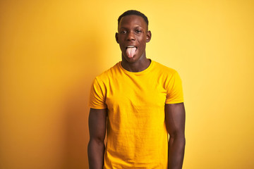 Young african american man wearing casual t-shirt standing over isolated yellow background sticking tongue out happy with funny expression. Emotion concept.