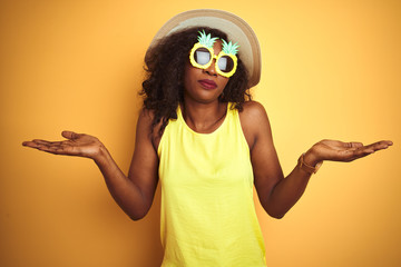 African american woman wearing funny pineapple sunglasses over isolated yellow background clueless...