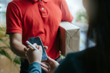 home delivery service man in red uniform and young woman customer appending signature in digital mobile phone receiving parcel post box from courier at home, express delivery, online shopping concept