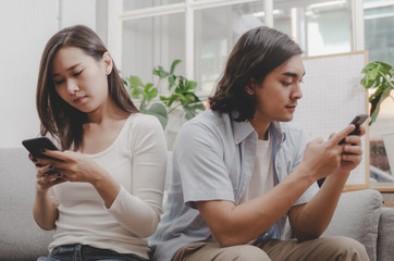 asian young couple man and woman absorbed and focused into mobile phone and sitting on sofa in living room at home, social media addiction, upset couple, family issues and relationship concept