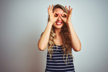 Young beautiful woman wearing stripes t-shirt standing over white isolated background doing ok gesture like binoculars sticking tongue out, eyes looking through fingers. Crazy expression.